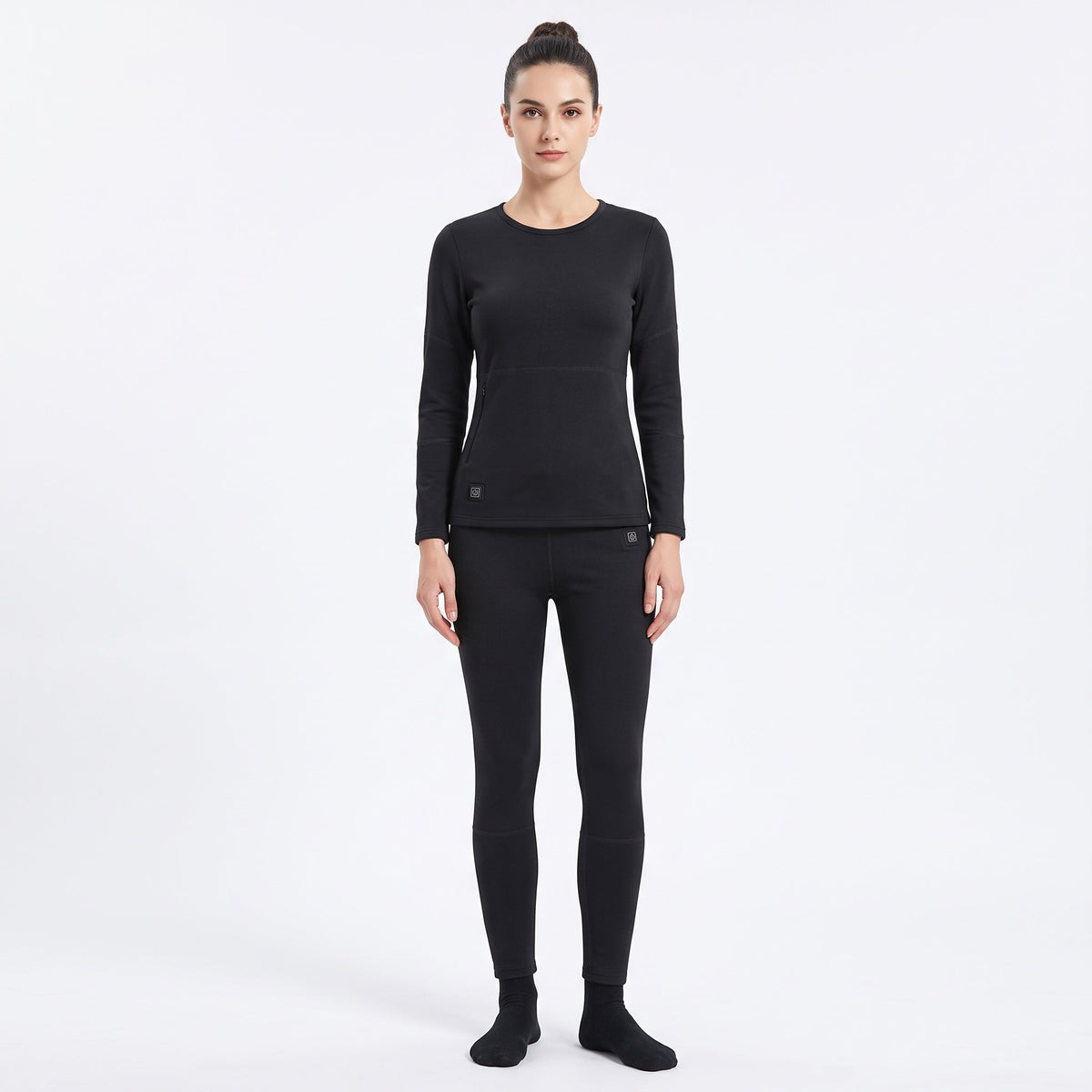 Full-body front view of a female model wearing a matching two-piece black smart heated thermal underwear set. The crew neck top has two black control icons at the hem, paired with full-length leggings against a solid white background.