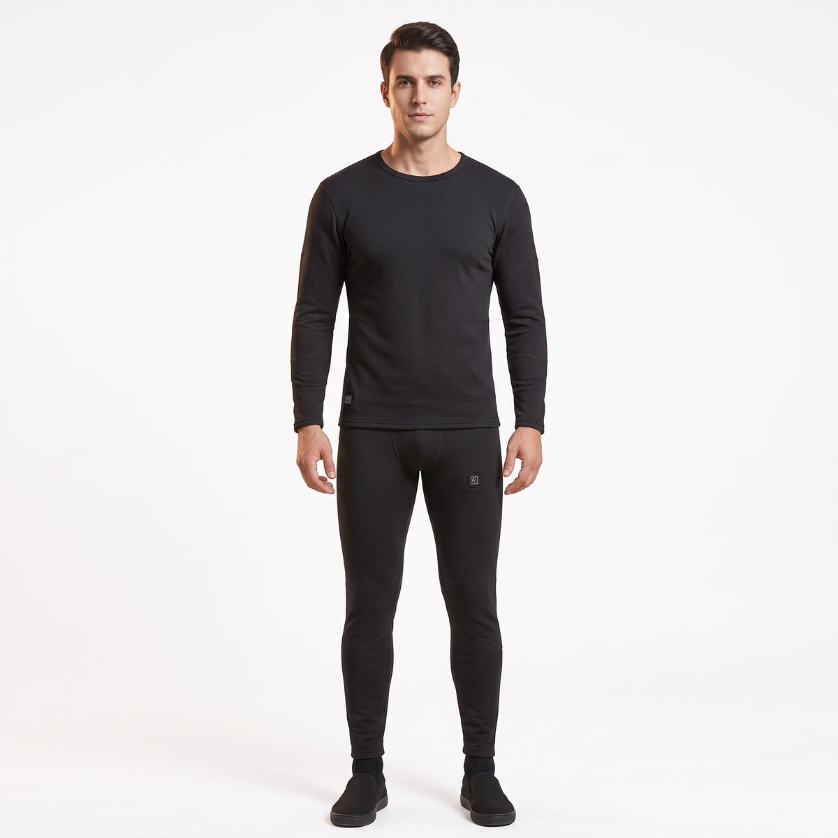 Full-body front view of a male model wearing a matching two-piece black smart heated thermal underwear set. The crew neck top has two black control icons at the hem, paired with full-length leggings against a solid white background.