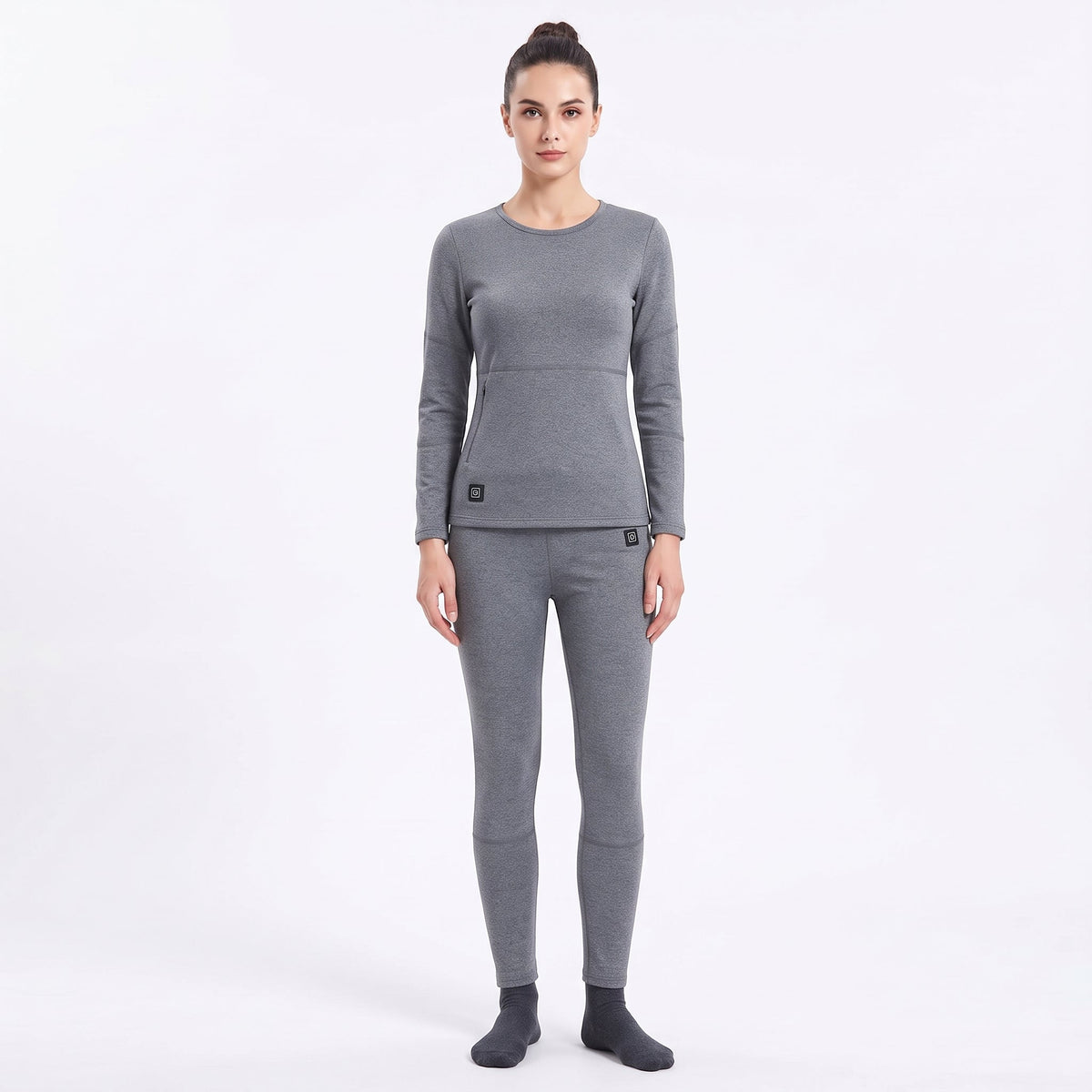 Full-body front view of a female model wearing a matching two-piece gray smart heated thermal underwear set. The crew neck top has two black control icons at the hem, paired with full-length leggings against a solid white background.